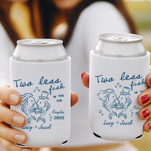 Puede incluir: Dos enfriadores de latas blancos con texto e ilustraciones azules. El texto dice "Two less fish in the sea" con una ilustración de una pareja de peces. Los nombres "Lucy + Jacob" también están impresos en los enfriadores. Los enfriadores están diseñados para una despedida de solteros.