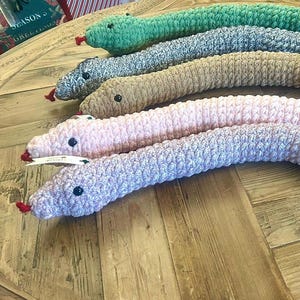 May include: A selection of handmade, plush snake toys in shades of green, grey, beige, pink, and lilac. Each snake has black button eyes and a red tongue. The toys are displayed on a wooden surface.