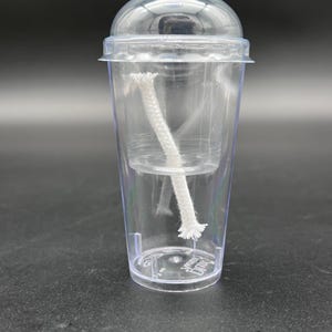 May include: A clear plastic cup with a dome lid and a white wick positioned inside. The cup features a transparent inner insert and a small white wick extending diagonally through the center. The base of the cup has embossed text visible.