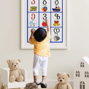 May include: A framed educational poster with Georgian alphabet letters and illustrations. A child in a yellow shirt and white pants points at the poster. Teddy bears, a wooden house, and a rainbow toy are in the foreground.