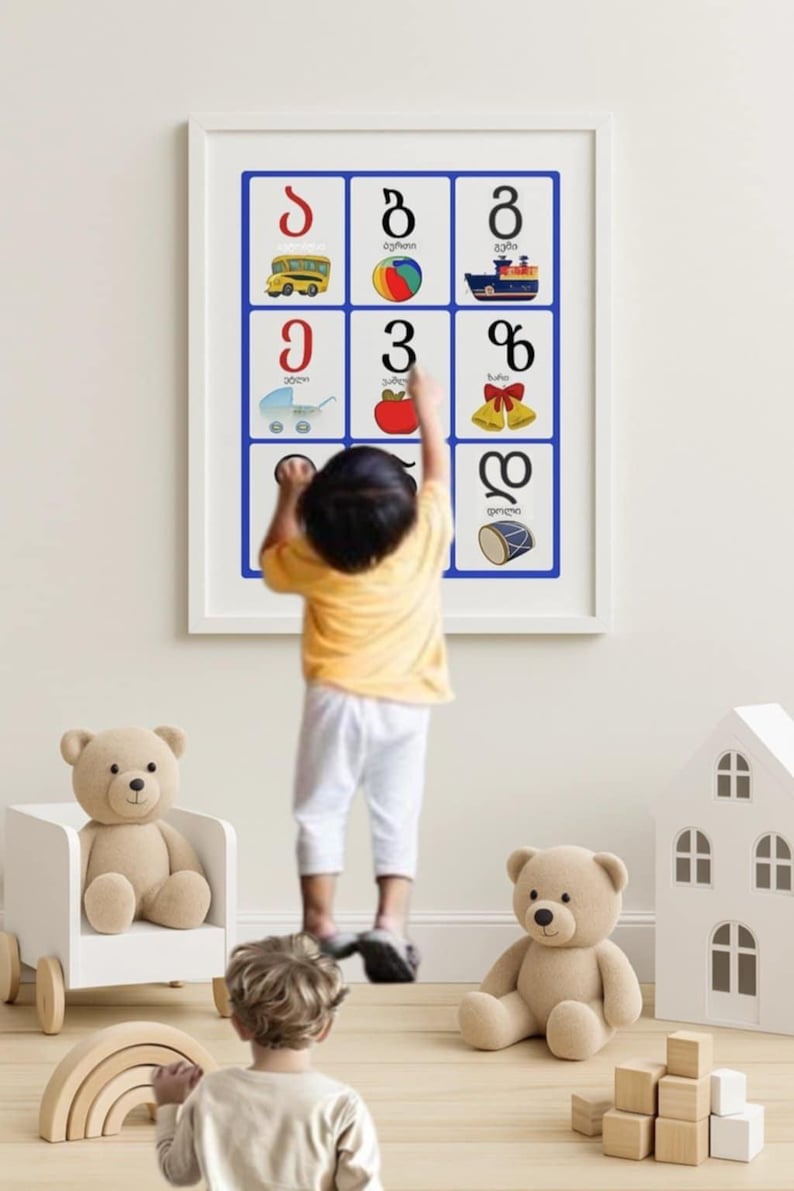May include: A framed educational poster with Georgian alphabet letters and illustrations. A child in a yellow shirt and white pants points at the poster. Teddy bears, a wooden house, and a rainbow toy are in the foreground.