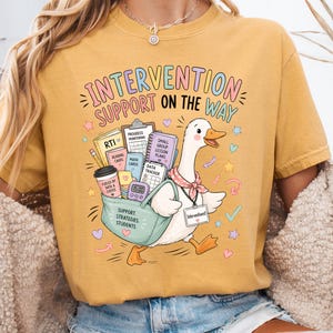 May include: A mustard yellow t-shirt featuring a graphic of a white duck wearing a pink neckerchief and an ID badge. The duck carries a teal bag filled with school supplies, including a coffee cup, books, and a clipboard. The text reads INTERVENTION SUPPORT ON THE WAY.