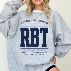 May include: A sport grey crewneck sweatshirt featuring dark blue text that reads REGISTERED BEHAVIOR TECHNICIAN, RBT, FOR EVERY STUDENT, EVERY SESSION, ABA THERAPY, BEHAVIOR SUPPORT, SCHOOL-BASED, and CLINICAL. The garment has long sleeves.