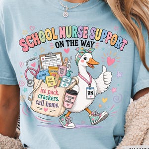 May include: A light blue t-shirt featuring a graphic of a white duck wearing a headband and an RN badge, carrying a bag filled with medical supplies. The text reads SCHOOL NURSE SUPPORT ON THE WAY, ice pack, crackers, call home, and caffeine for compassion.
