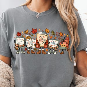 May include: A charcoal grey t-shirt featuring a graphic print of five anthropomorphic school supplies with smiling faces, wearing boots and scarves, surrounded by autumn leaves. The characters hold items labeled CAT, 2+3, and MAP 4+2.