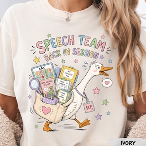 May include: An ivory t-shirt featuring a colorful graphic of a goose carrying a bag filled with speech therapy supplies, including an AAC device, articulation cards, and data sheets. The text reads SPEECH TEAM BACK IN SESSION with stars.