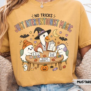 May include: A mustard yellow t-shirt featuring a graphic of a duck wearing a witch hat, sitting at a table with two ghosts. The text reads NO TRICKS JUST INTERVENTIONIST MAGIC. The illustration includes a clipboard, math problems, and pumpkins.