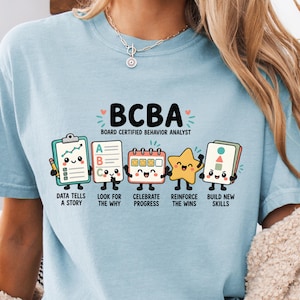 May include: A light blue t-shirt featuring the text BCBA BOARD CERTIFIED BEHAVIOR ANALYST and five cartoon characters representing a clipboard, a list, a calendar, a star, and flashcards with the phrases DATA TELLS A STORY, LOOK FOR THE WHY, CELEBRATE PROGRESS, REINFORCE THE WINS, and BUILD NEW SKILLS.