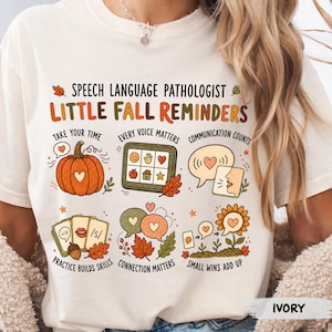 May include: An ivory t-shirt featuring the text SPEECH LANGUAGE PATHOLOGIST LITTLE FALL REMINDERS and six illustrations including a pumpkin, a communication tablet, speech bubbles, flashcards, autumn leaves, and a sunflower with heart icons.