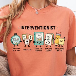 May include: A terracotta-colored t-shirt featuring the text INTERVENTIONIST and cartoon illustrations of a stopwatch, letter blocks spelling bat, a data binder, a book, a pencil, and a sticky note with the phrases One Step At A Time, Build It Up, Track The Growth, Practice Counts, and Small Wins Matter.