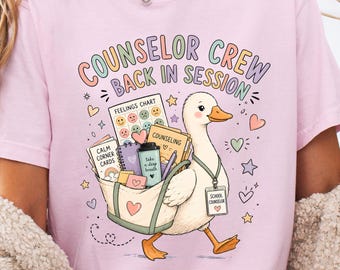 May include: A light pink t-shirt featuring a graphic of a white duck carrying a tote bag filled with school supplies, including a feelings chart, calm corner cards, and a coffee cup. The text reads COUNSELOR CREW BACK IN SESSION and SCHOOL COUNSELOR.