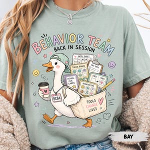 May include: A sage green t-shirt featuring a cartoon duck wearing a baseball cap and a BCBA badge, holding a coffee cup and a tote bag filled with educational materials. The text reads BEHAVIOR TEAM BACK IN SESSION and TOOLS CHANGE LIVES.