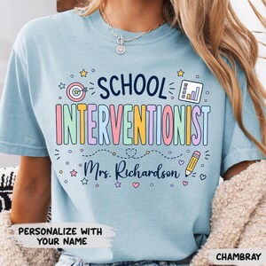 May include: A light blue t-shirt featuring the text "SCHOOL INTERVENTIONIST" in colorful letters, along with a target, a chart, a pencil, and the name "Mrs. Richardson" printed in dark blue. The design includes small stars and hearts.
