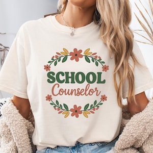 Könnte beinhalten: Cremefarbenes T-Shirt mit dem Schriftzug "SCHOOL Counselor" in Grün und Koralle, umgeben von einem floralen Kranz. Der Kranz zeigt orangefarbene Blüten und grüne Blätter. Das Shirt hat eine lockere Passform.