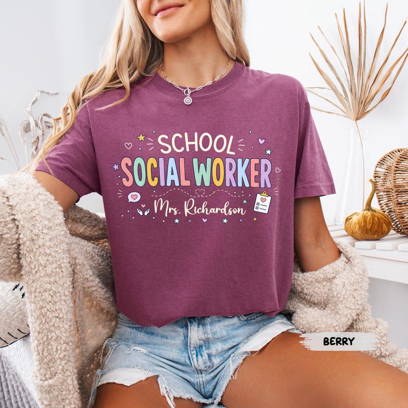 K&ouml;nnte beinhalten: Ein beerenfarbenes Kurzarm-T-Shirt mit dem Text "SCHOOL SOCIAL WORKER" in bunten Buchstaben, dem Namen "Mrs. Richardson" in wei&szlig;er Schreibschrift, einem Klemmbrett-Symbol sowie kleinen dekorativen Herzen und Sternen.
