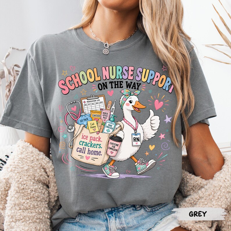May include: A grey t-shirt featuring a graphic of a white duck wearing a headband and an RN badge. The duck carries a tote bag filled with medical supplies and a coffee cup. The text reads "SCHOOL NURSE SUPPORT ON THE WAY" and "ice pack, crackers, call home."