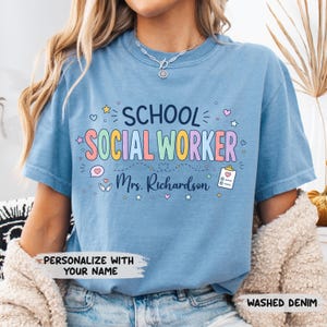 K&ouml;nnte beinhalten: Ein verwaschenes, denimblaues T-Shirt mit dem Text SCHOOL SOCIAL WORKER in bunten Buchstaben und dem Namen Mrs. Richardson darunter. Das Design enth&auml;lt kleine grafische Symbole wie Sterne, Herzen, eine Sprechblase und ein Klemmbrett mit Herz.