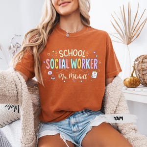 K&ouml;nnte beinhalten: Ein rostorangefarbenes T-Shirt mit dem Text "SCHOOL SOCIAL WORKER" in bunten Buchstaben und "Mrs. Mitchell" darunter. Das Design enth&auml;lt kleine grafische Symbole wie Herzen, Sterne, ein Klemmbrett und H&auml;nde, die auf die Vorderseite gedruckt sind.