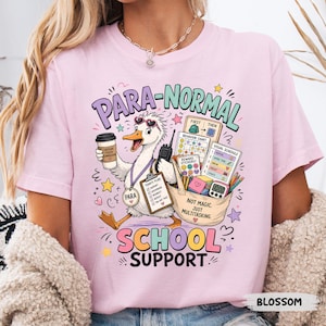 May include: A pale pink t-shirt with the text "PARA-NORMAL SCHOOL SUPPORT" and a cartoon goose holding a coffee cup and a walkie-talkie. The goose wears a "PARA" lanyard. The shirt also features a tote bag with school supplies and the words "NOT MAGIC. JUST MULTITASKING."