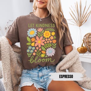 Puede incluir: Una camiseta marr&oacute;n con un dise&ntilde;o floral y el texto "Let Kindness Bloom" en letras marrones. La camiseta la lleva puesta una persona con el pelo rubio. El texto "Espresso" est&aacute; escrito en blanco sobre un fondo marr&oacute;n en la esquina inferior derecha de la imagen.