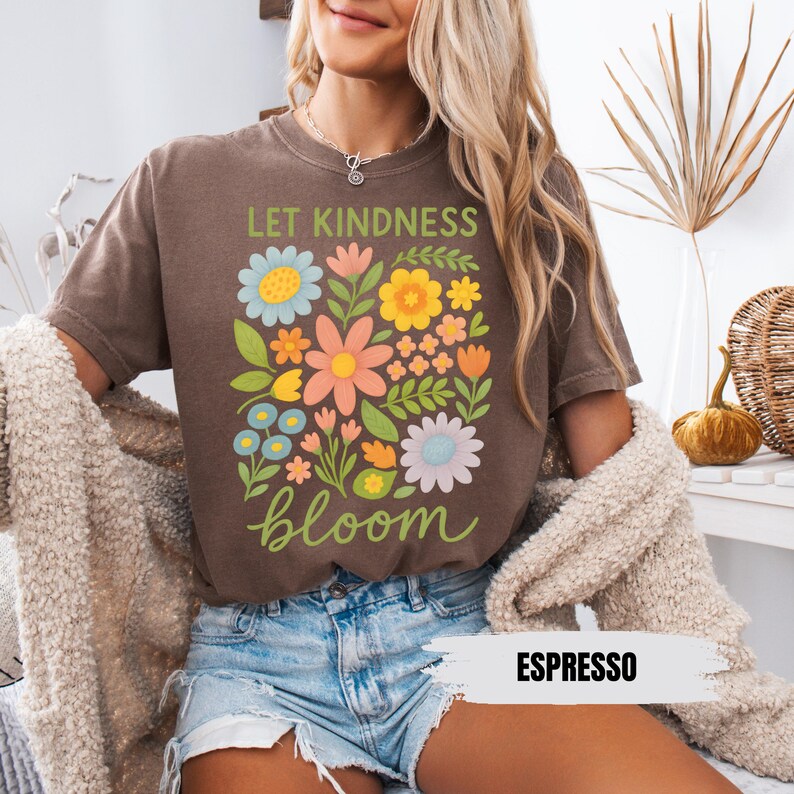 Puede incluir: Una camiseta marr&oacute;n con un dise&ntilde;o floral y el texto "Let Kindness Bloom" en letras marrones. La camiseta la lleva puesta una persona con el pelo rubio. El texto "Espresso" est&aacute; escrito en blanco sobre un fondo marr&oacute;n en la esquina inferior derecha de la imagen.