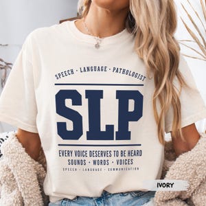 May include: An ivory short-sleeved t-shirt featuring dark blue text that reads SPEECH LANGUAGE PATHOLOGIST, SLP, EVERY VOICE DESERVES TO BE HEARD, SOUNDS WORDS VOICES, and SPEECH LANGUAGE COMMUNICATION in a collegiate-style graphic design.