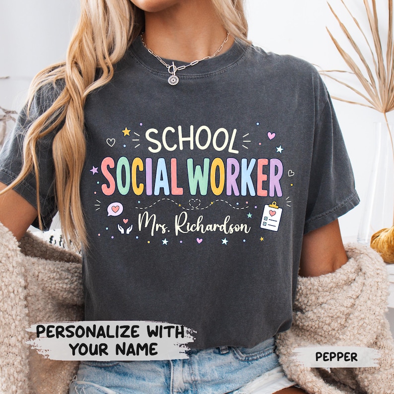 K&ouml;nnte beinhalten: Ein anthrazitfarbenes T-Shirt mit dem Text SCHOOL SOCIAL WORKER in bunten Buchstaben und dem Namen Mrs. Richardson darunter. Das Design enth&auml;lt kleine Symbole wie Herzen, Sterne und ein Klemmbrett sowie den Text PERSONALIZE WITH YOUR NAME.