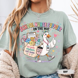 May include: A sage green t-shirt featuring a graphic of a white duck wearing a headband and an RN badge, carrying a tote bag filled with medical supplies. The text reads "SCHOOL NURSE SUPPORT ON THE WAY" and "ice pack, crackers, call home."