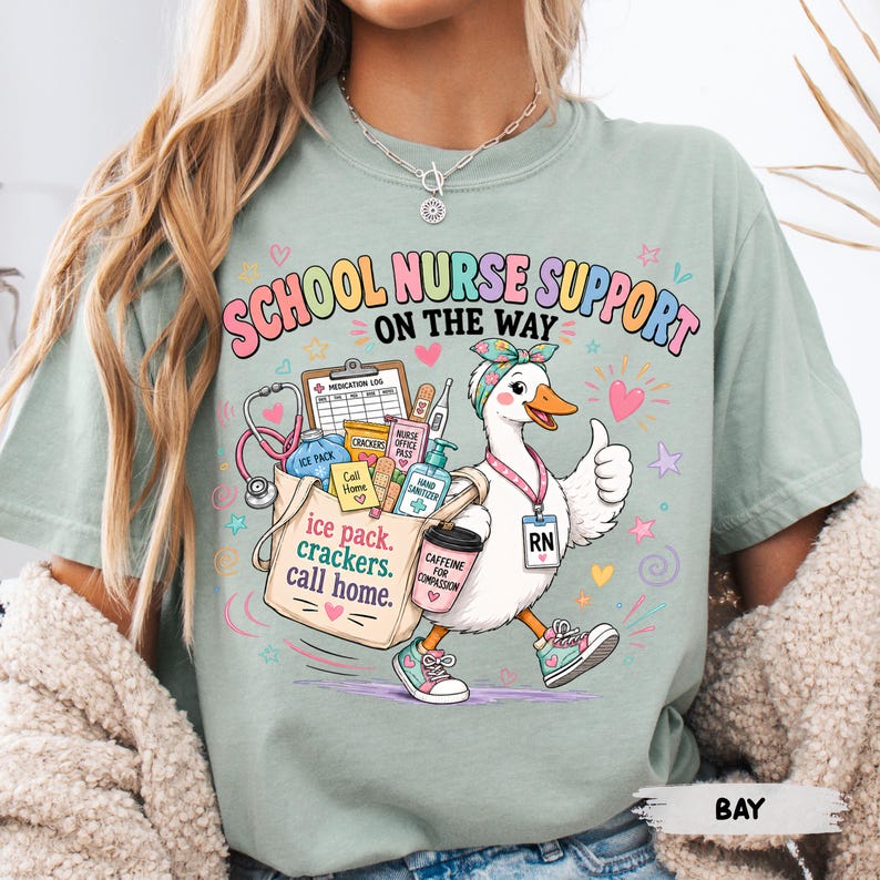 May include: A sage green t-shirt featuring a graphic of a white duck wearing a headband and an RN badge, carrying a tote bag filled with medical supplies. The text reads "SCHOOL NURSE SUPPORT ON THE WAY" and "ice pack, crackers, call home."