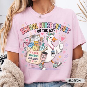May include: A light pink t-shirt featuring a graphic of a white duck wearing a headband and sneakers, carrying a tote bag filled with medical supplies. The text reads SCHOOL NURSE SUPPORT ON THE WAY, ice pack, crackers, call home, and CAFFEINE FOR COMPASSION.