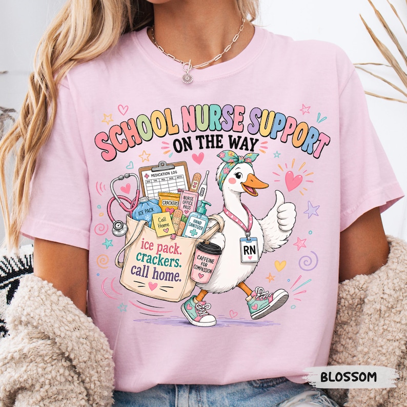 May include: A light pink t-shirt featuring a graphic of a white duck wearing a headband and sneakers, carrying a tote bag filled with medical supplies. The text reads SCHOOL NURSE SUPPORT ON THE WAY, ice pack, crackers, call home, and CAFFEINE FOR COMPASSION.