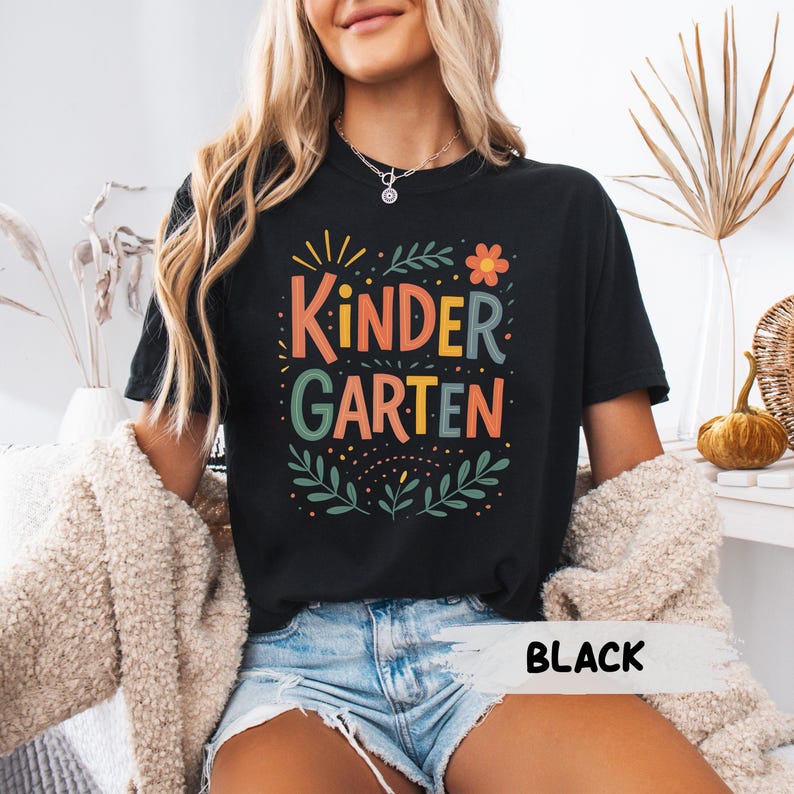 Może przedstawiać: Czarny t-shirt z napisem "Kindergarten" w kolorowych literach. Projekt zawiera kwiatowe i liściaste akcenty. Słowo "BLACK" jest wydrukowane na dole koszulki.