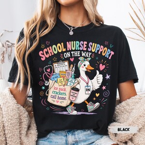 May include: A black t-shirt featuring a graphic of a white duck wearing a headband and sneakers, carrying a bag filled with medical supplies. The text reads SCHOOL NURSE SUPPORT ON THE WAY, ice pack, crackers, call home, and CAFFEINE FOR COMPASSION.