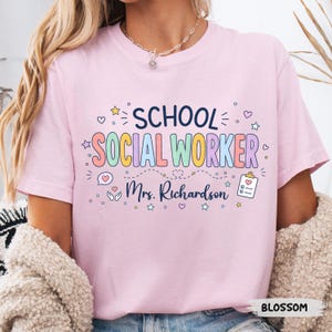 K&ouml;nnte beinhalten: Ein hellrosa T-Shirt mit kurzen &Auml;rmeln, das den dunkelblauen Schriftzug SCHOOL und den bunten Schriftzug SOCIAL WORKER zeigt. Darunter ist der Name Mrs. Richardson in dunkelblauer Schreibschrift gedruckt, erg&auml;nzt durch kleine Herz-, Klemmbrett- und Sternsymbole.