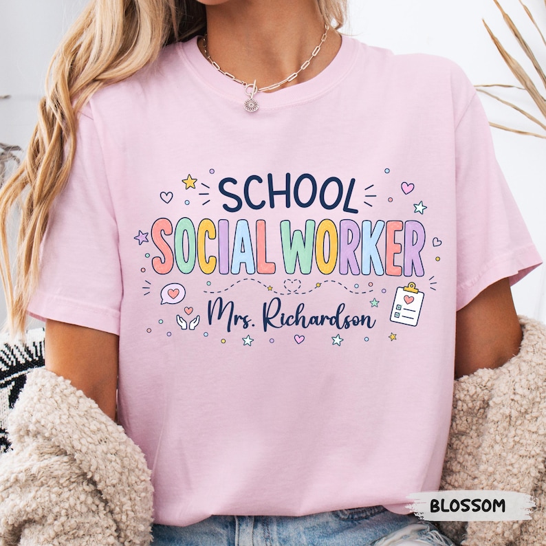 K&ouml;nnte beinhalten: Ein hellrosa T-Shirt mit kurzen &Auml;rmeln, das den dunkelblauen Schriftzug SCHOOL und den bunten Schriftzug SOCIAL WORKER zeigt. Darunter ist der Name Mrs. Richardson in dunkelblauer Schreibschrift gedruckt, erg&auml;nzt durch kleine Herz-, Klemmbrett- und Sternsymbole.