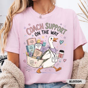 May include: Pale pink t-shirt featuring a cartoon goose with a tote bag and coffee cup. The text on the shirt says "COACH SUPPORT ON THE WAY." The tote bag contains school supplies. Other text includes "Lesson Planner" and "Instructional Coach."