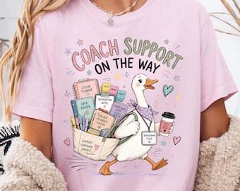 May include: Pink t-shirt with a graphic of a goose carrying a tote bag filled with school supplies and a coffee cup. Text reads "COACH SUPPORT ON THE WAY." Additional text includes "Lesson Planner," "Data Binder," and "Instructional Coach."