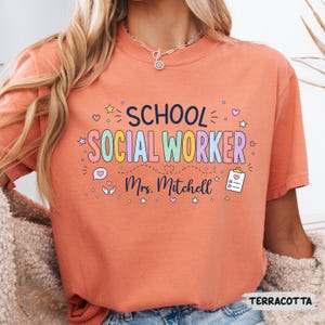 K&ouml;nnte beinhalten: Ein terrakottafarbenes T-Shirt mit dem dunkelblauen Schriftzug SCHOOL und dem bunten Schriftzug SOCIAL WORKER. Darunter ist der Name Mrs. Mitchell in dunkelblauer Schreibschrift gedruckt, erg&auml;nzt durch Symbole einer Sprechblase, H&auml;nden und eines Klemmbretts.