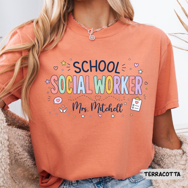 K&ouml;nnte beinhalten: Ein terrakottafarbenes T-Shirt mit dem dunkelblauen Schriftzug SCHOOL und dem bunten Schriftzug SOCIAL WORKER. Darunter ist der Name Mrs. Mitchell in dunkelblauer Schreibschrift gedruckt, erg&auml;nzt durch Symbole einer Sprechblase, H&auml;nden und eines Klemmbretts.