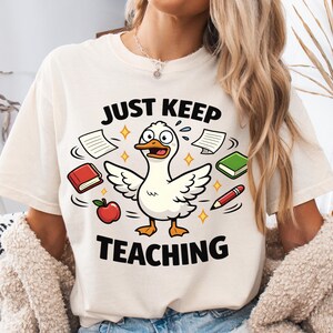 May include: Cream-colored t-shirt with a cartoon duck design. The duck is surrounded by books, an apple, and a pencil. The text "JUST KEEP TEACHING" is printed above and below the duck illustration.