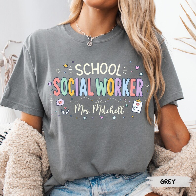K&ouml;nnte beinhalten: Ein anthrazitfarbenes T-Shirt mit dem Text "SCHOOL SOCIAL WORKER" in bunten Pastellbuchstaben und dem Namen "Mrs. Mitchell" darunter. Das Design enth&auml;lt kleine grafische Symbole wie Sterne, Herzen, eine Sprechblase und ein Klemmbrett.