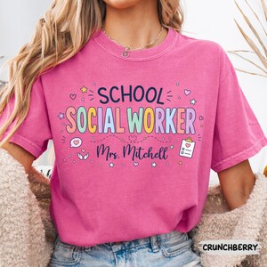 K&ouml;nnte beinhalten: Ein leuchtend pinkfarbenes T-Shirt mit dem Text "SCHOOL SOCIAL WORKER" in bunten Buchstaben und "Mrs. Mitchell" darunter. Das Design enth&auml;lt kleine Symbole wie Sterne, Herzen, eine Sprechblase mit einem Herz und ein Klemmbrett mit einem Herz.