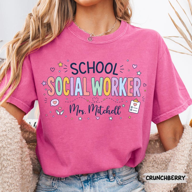 K&ouml;nnte beinhalten: Ein leuchtend pinkfarbenes T-Shirt mit dem Text "SCHOOL SOCIAL WORKER" in bunten Buchstaben und "Mrs. Mitchell" darunter. Das Design enth&auml;lt kleine Symbole wie Sterne, Herzen, eine Sprechblase mit einem Herz und ein Klemmbrett mit einem Herz.