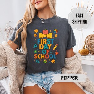 Könnte beinhalten: Dunkelgraues T-Shirt mit dem Text "FIRST DAY OF SCHOOL" in bunten Buchstaben. Das Design umfasst einen Apfel, eine Schleife, einen Bleistift und andere schulbezogene Grafiken. Das Shirt ist ein bequemer, lässiger Stil.