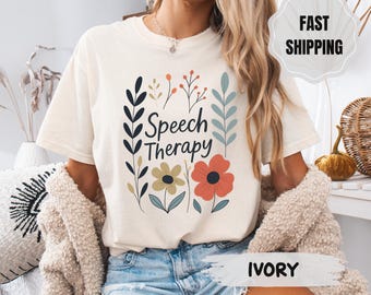 May include: Ivory-colored t-shirt with the words "Speech Therapy" in black script, surrounded by floral illustrations in navy, green, orange, and light blue. The shirt has a relaxed fit and short sleeves.