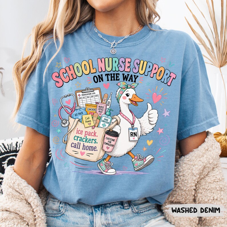May include: A light blue t-shirt featuring a graphic of a white duck wearing a headband, sneakers, and an RN badge. The duck holds a tote bag filled with medical supplies and a coffee cup. Text reads "SCHOOL NURSE SUPPORT ON THE WAY".