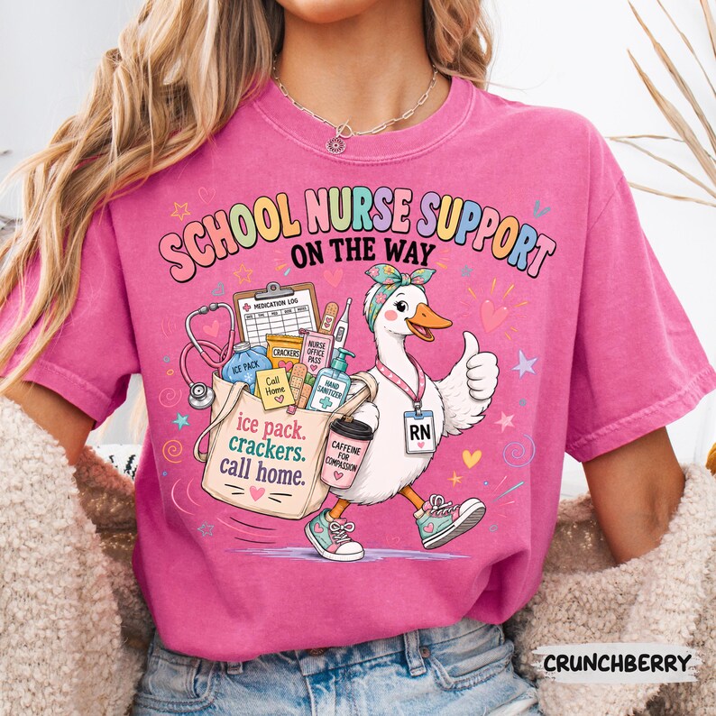 May include: A pink t-shirt featuring a graphic of a duck wearing a headband and an RN badge, giving a thumbs up. The text reads SCHOOL NURSE SUPPORT ON THE WAY and ice pack, crackers, call home. The duck carries a bag with medical supplies.