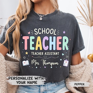 May include: A charcoal grey t-shirt featuring the text SCHOOL TEACHER and TEACHER ASSISTANT in colourful pastel letters. Below, the name Mrs. Thompson is printed in white script, accompanied by small icons of a book, a clipboard, and stars.