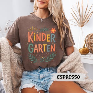 Może przedstawiać: Koszulka w kolorze espresso z napisem "KINDERGARTEN" w kolorowej, zabawnej czcionce. Projekt zawiera kwiatowe i liściaste akcenty. Koszulka jest połączona z dżinsowymi szortami.