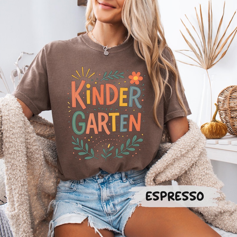Może przedstawiać: Koszulka w kolorze espresso z napisem "KINDERGARTEN" w kolorowej, zabawnej czcionce. Projekt zawiera kwiatowe i liściaste akcenty. Koszulka jest połączona z dżinsowymi szortami.